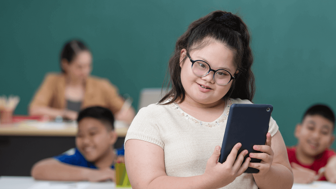 a young girl who has Downs Syndrome is stood in front of a class full of students. She is smiling and tilting her head to one side whilst wearing glasses and holding a tablet