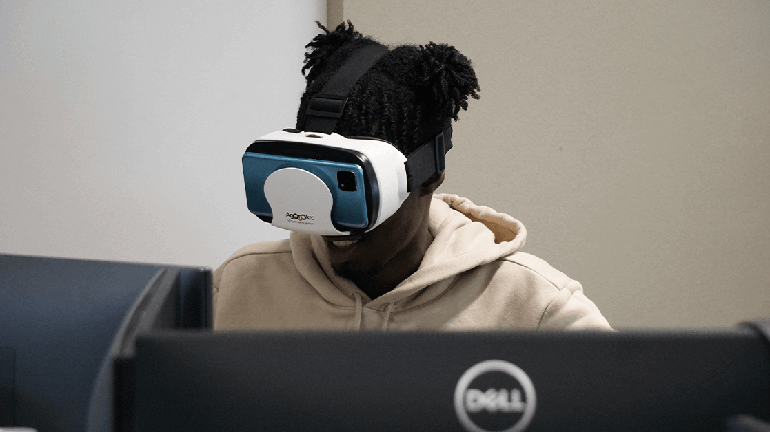 A Black student sat smiling, whilst wearing a VR headset at a computer