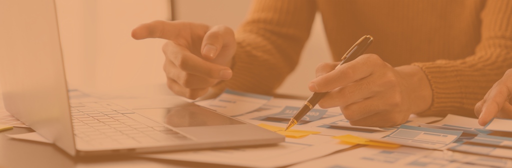 two people are sat at a desk looking at a laptop and using their audit findings to adapt their wireframe design, which are spread out across the desk.