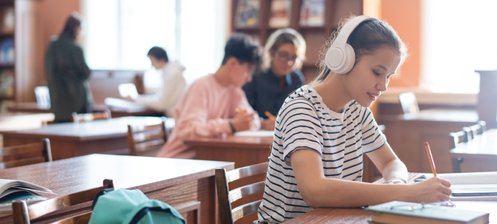A secondary school student, who is a girl with tied back brown hair and is wearing a white and blue striped t-shirt, is sat wearing white headphones whilst taking notes within a classroom