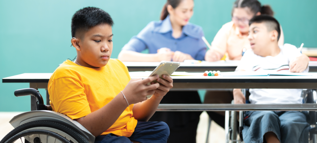 A secondary school student, who is Asian and a wheelchair user, has short black hair and is wearing a bright yellow t-shirt. He is in a classroom and is using a silver tablet to access learning resources.