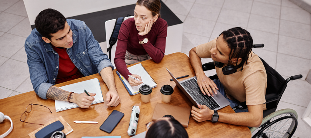 four university students are sat around a table talking. Three of them are taking notes in a pad, whilst a Black wheelchair user, who has headphones on around his neck, is using a laptop instead