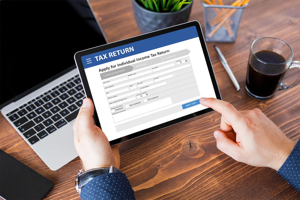 Someone is a navy shirt is sat at a wooden desk holding a tablet. Displayed on the screen is an online form to complete a tax return.