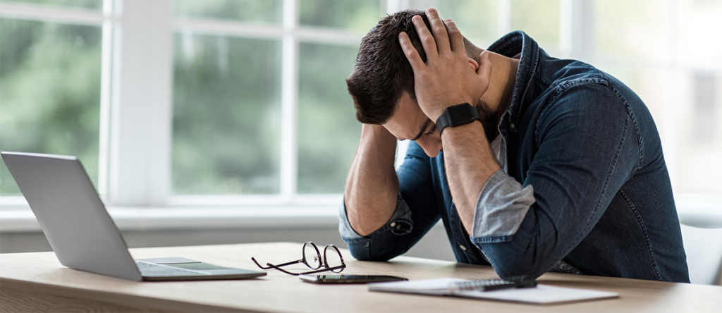 A man, who has short brown hair and is wearing a denim shirt, is sat with his head held in his hands in despair at a desk in front of a laptop