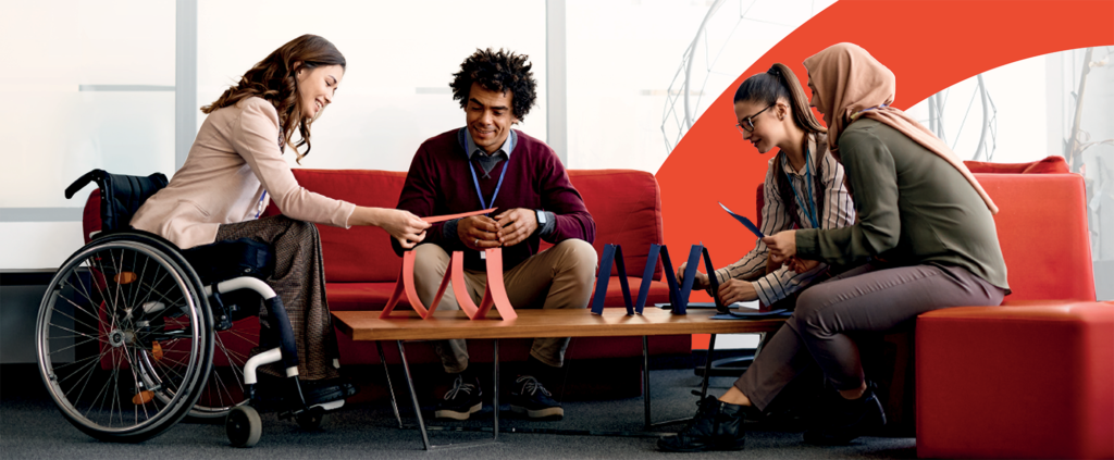 Four office workers, one of which is a female wheelchair user, are sat happily on sofas playing a game stacking a deck of cards.