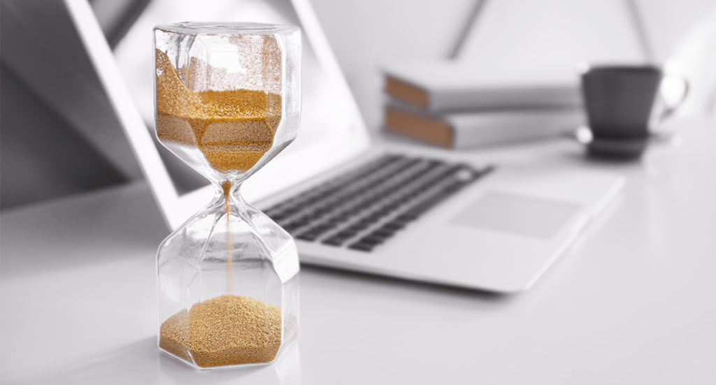 A silver laptop is on a white desk. Next to it is a hourglass timer.