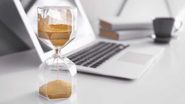 A silver laptop is on a white desk. Next to it is a hourglass timer.