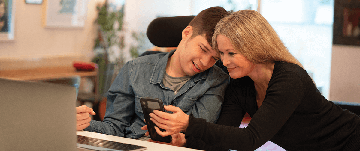 A mother is leaning over her son at a desk, who has Cerebral Palsy. She is assisting him accessing the web with a mobile device.