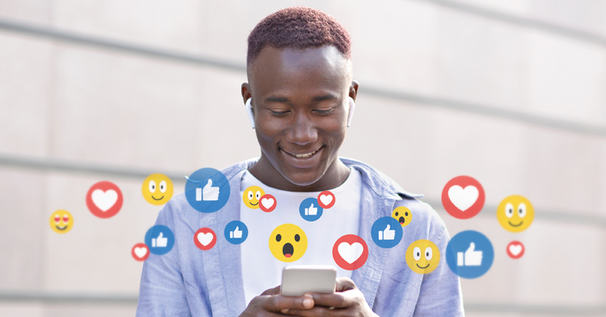 A young Black man, in a pale blue hooded jacket and wearing white headphones, is stood outdoors smiling at his mobile phone. Emerging from the phone are multiple social media likes, loves, and emojis.